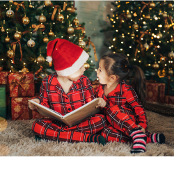 Deux enfants qui lisent un livre assis sur le sol devant deux arbres de noël en plein décor.