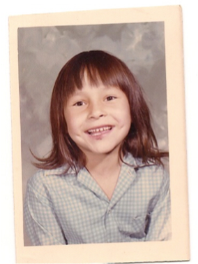 Portrait scolaire de Phyllis Webstad en tant que jeune fille. Elle a les cheveux bruns aux épaules avec une frange et une chemise grise. Elle sourit.