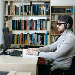 Un homme portant des écouteurs et des lunettes est assis à un bureau face à un ordinateur et lit du braille. Il y a des étagères de livres en arrière-plan.
