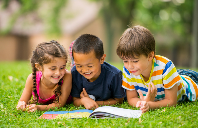 Three kids lying on the grass looking at a book. 
